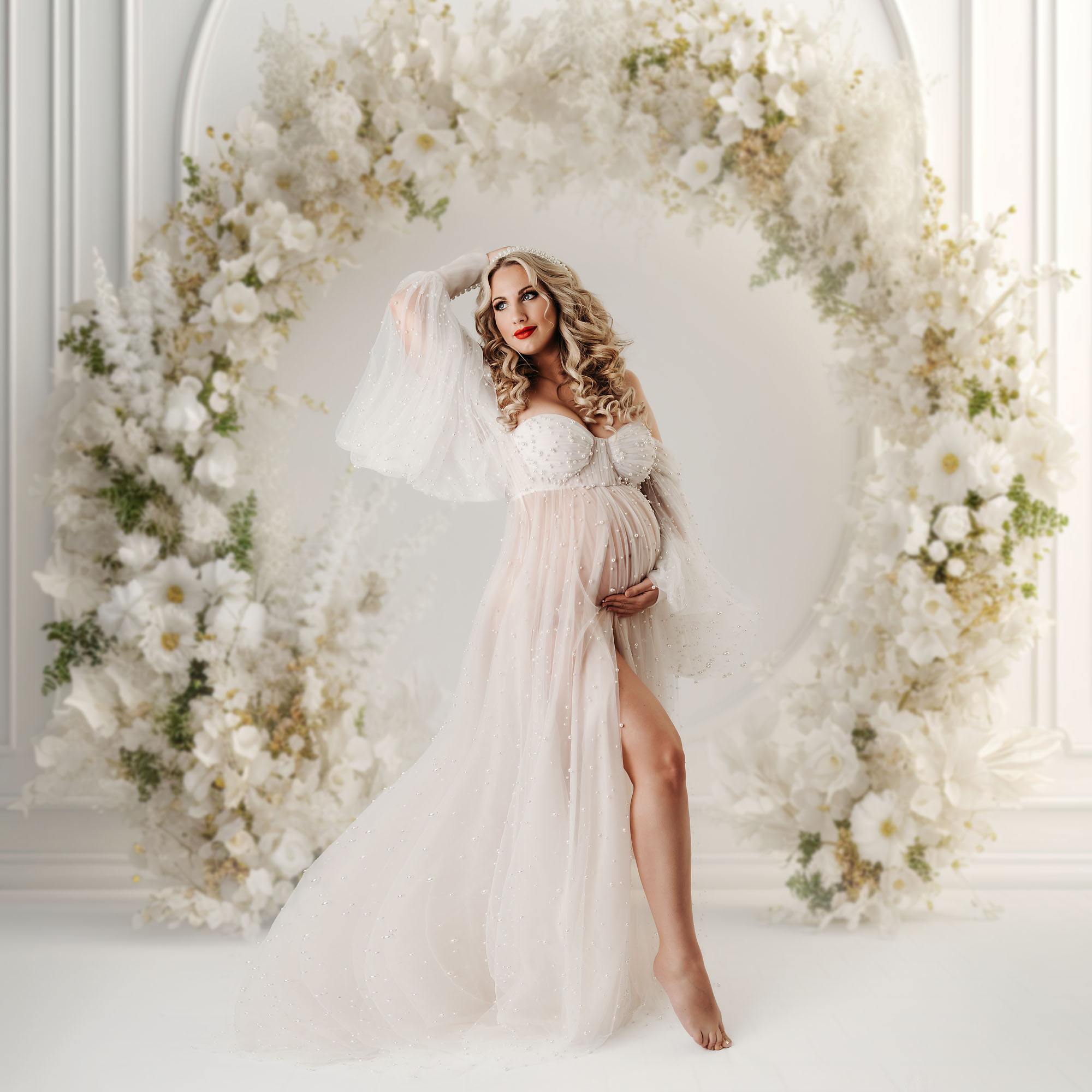 Schwangere Frau im langen Kleid mit vielen Perlen und runden Blumenkranz im Hintergrund